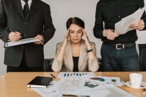 Lady sat down looking stressed with her hands held to her face, next to two men with lots of documents on the table